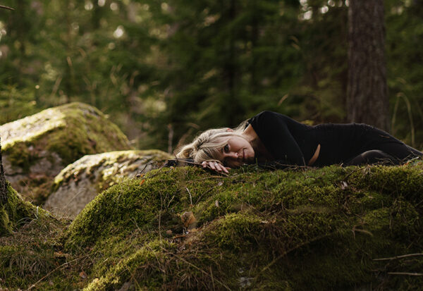 Woman resting on mossy ground in a forest, lying on her side with her head on her arm in a black outfit.